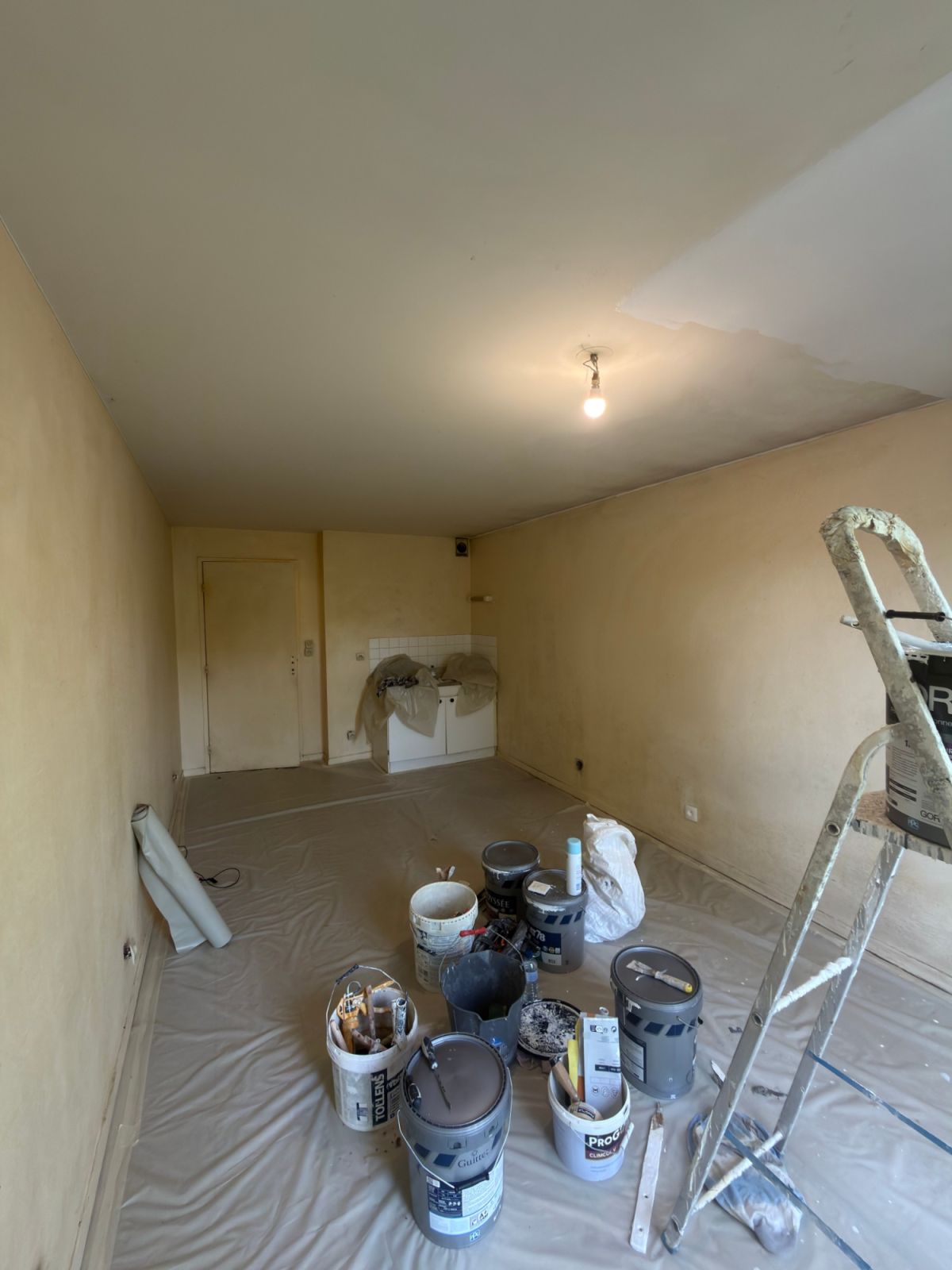 Studio avant rénovation, murs jaunis, sol bâché et pots de peinture - chantier Yvelines